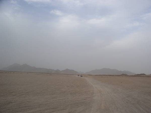 Vehicle on sandy desert track, dust-hazed mountains under grey overcast sky