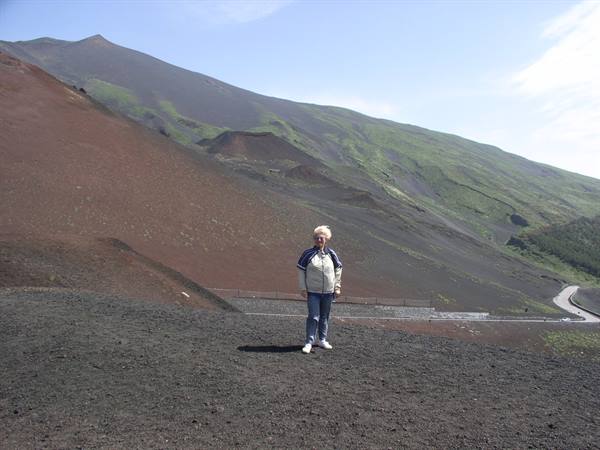 Woman standing on dark volcanic sand beneath Etna's green craters