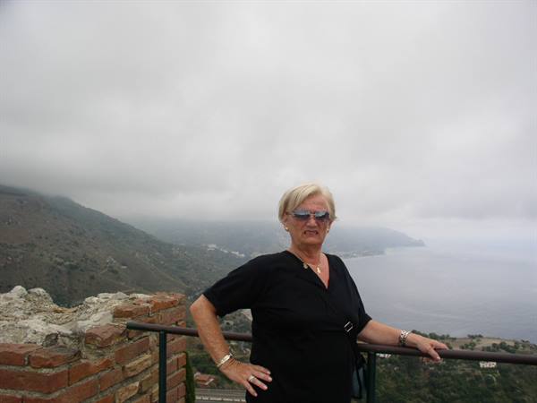 Blonde woman in sunglasses leaning on a railing by brick walls, coast and sea under fog