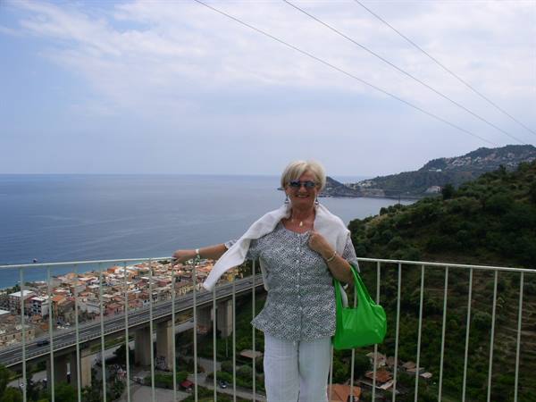 Blonde woman with green bag leaning on railing above Sicilian coastline