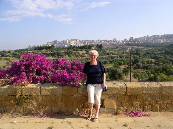 Woman leaning on a stone wall beside pink flowers, hilltop town behind