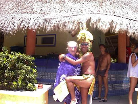 Tourist hugs a man in Maya costume with green headdress under thatched palapa