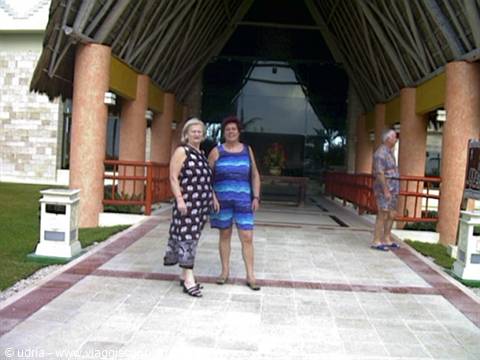 Two women in summer dresses smile under a thatched-roof pavilion entrance