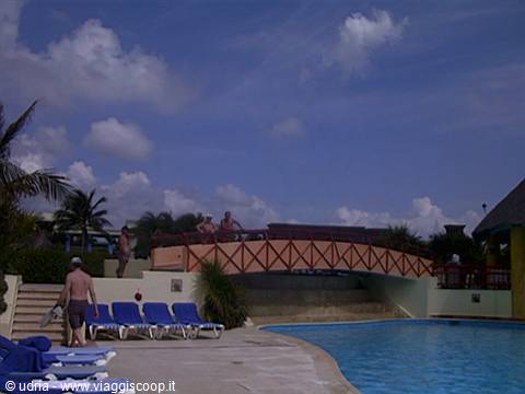 Orange wooden bridge over the pool, blue loungers and tourists under cloudy sky