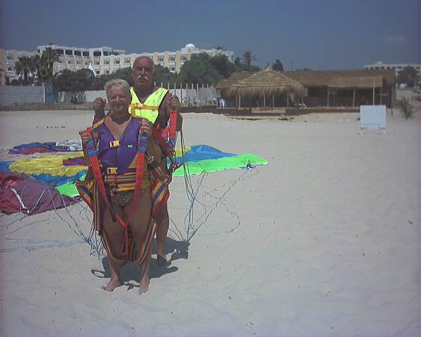 Couple in parasailing harness on sand, colorful sail and thatched hut behind