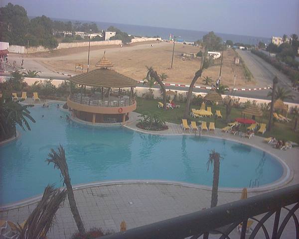 Blue pool with round thatched bar, yellow loungers, palm trees and sea beyond