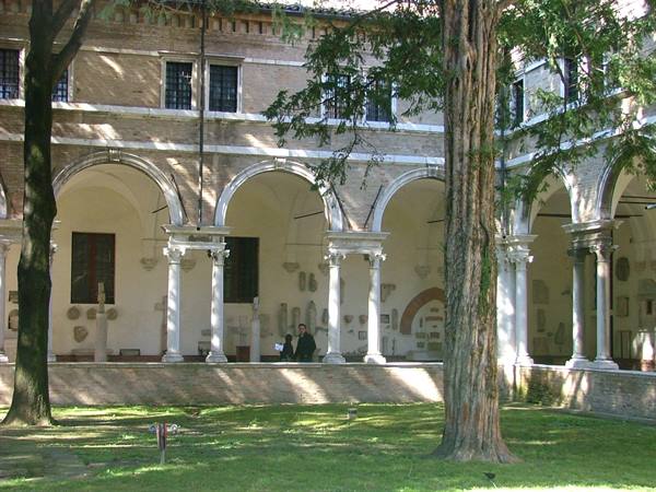 Cloister with brick arches, Roman columns and stone inscriptions, sunlight through trees