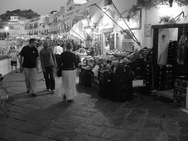 Evening cobblestone street with passersby and illuminated fruit stalls, in black and white
