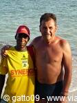 Two smiling men on the beach, one in yellow Synesis shirt, sea and sand