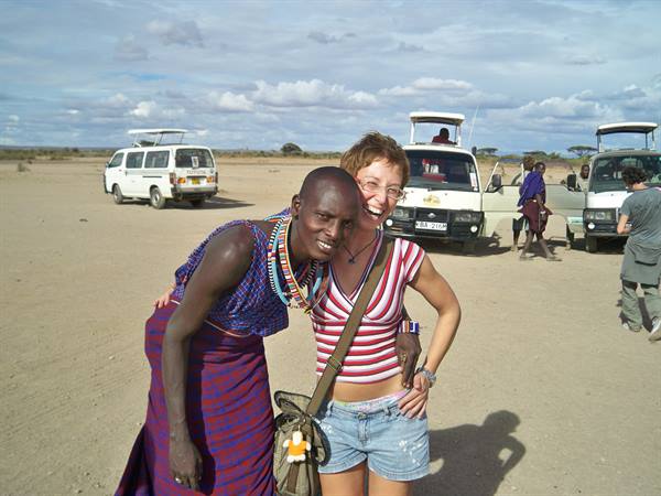 Maasai woman in beaded necklaces hugs a smiling tourist by safari vans