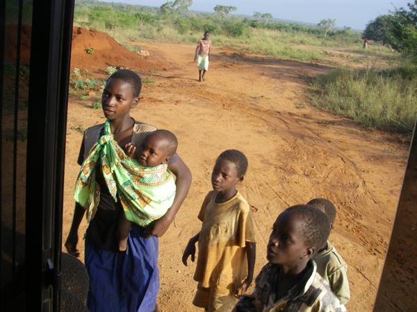 Woman carrying a baby wrapped in green cloth beside children on red dirt road