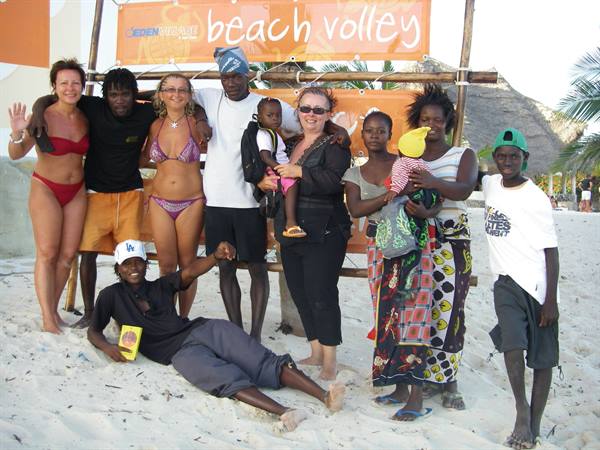 Tourists and locals pose on the sand in front of the beach volleyball sign