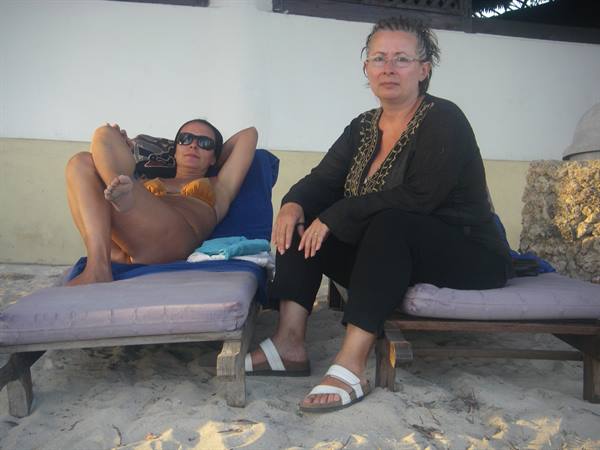 Two women on wooden loungers on the sand, one sunbathing wearing sunglasses