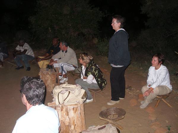 Tourists sitting on wooden stools around tree-trunk tables at night, safari camp