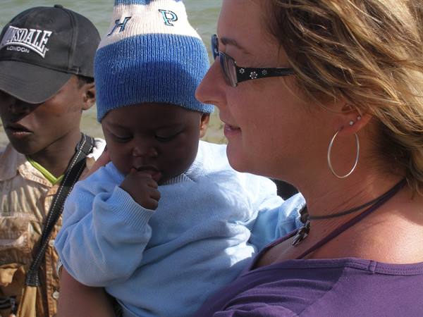Blonde woman in sunglasses holding a baby in a blue knit hat by the water