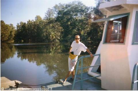 Man in white t-shirt and yellow hat standing on riverboat among trees