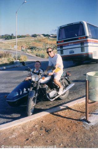 Two men on a motorcycle with blue sidecar beside a bus