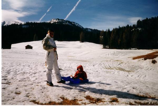 Child on blue sled in the snow, woman in white suit beside, mountains and firs