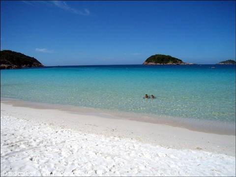 White sand beach, two people swimming in turquoise water, green islets in background