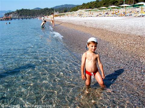 Child in red swimsuit and white cap on pebble beach with umbrellas and clear water