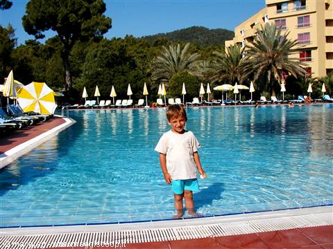 Child standing in the pool among yellow umbrellas, palms and hotel in sunlight