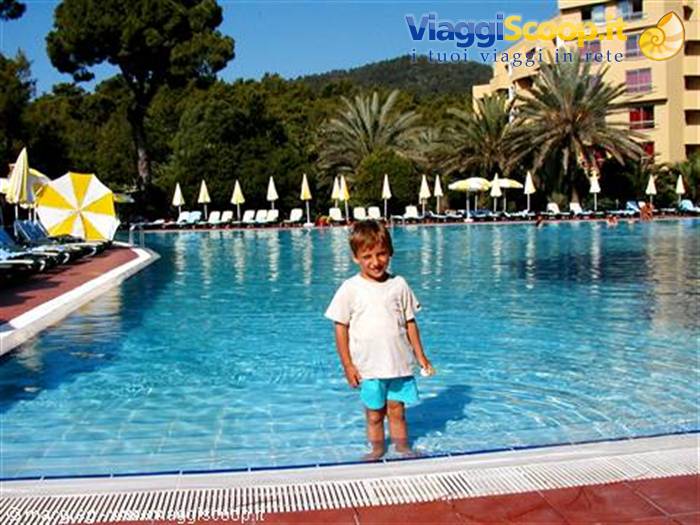 Child standing in the pool among yellow umbrellas, palms and hotel in sunlight