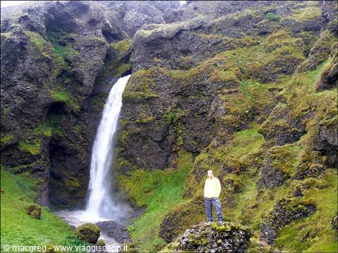 Man in yellow jacket on mossy rock in front of waterfall between green cliffs