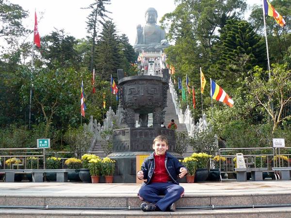 Boy sitting on steps in front of a giant bronze Buddha statue, colorful flags, daylight