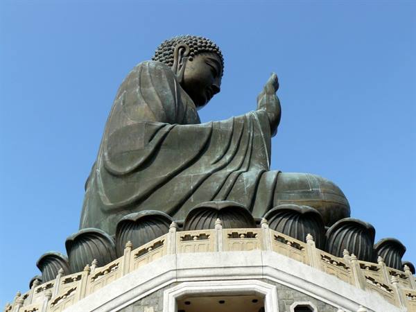 Large bronze seated Buddha statue with raised hand, blue sky, sunlight