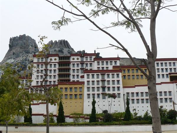 White and ochre multi-story building beneath gray rocks, framed by trees