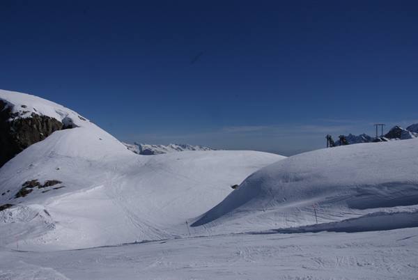 Snowy ski slopes between rocks under blue sky, ski lift in the distance