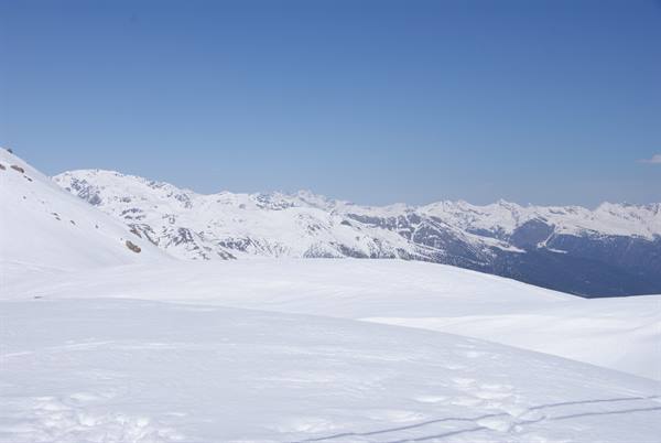 Snowy ski slope with tracks, snow-covered alpine peaks under clear blue sky