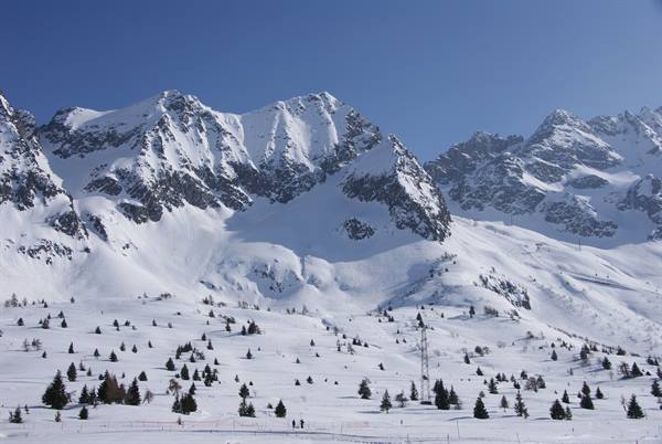 Snow-covered mountains under blue sky, scattered firs, two people walking on snow