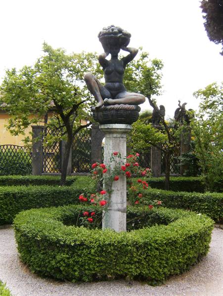 Female stone statue on column, surrounded by hedges and red roses in garden