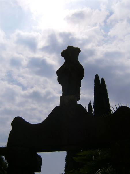 Stone statue silhouetted against clouds, cypress and foliage in background