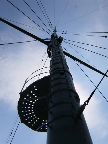 Ship's metal mast with perforated crow's nest and rigging against the sky