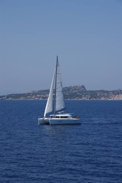 Sailing catamaran cruising on blue sea, rocky coastline behind