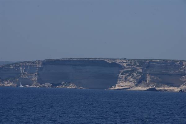 Limestone cliffs plunging into blue sea, a white sailboat in the distance at Lampedusa