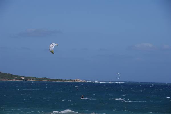 Kitesurfer with white sail glides on blue sea near green rocky coast