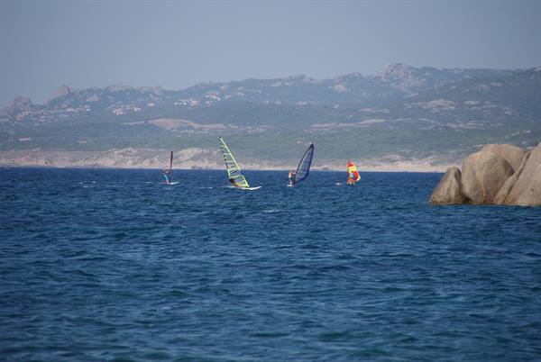 Four windsurfers gliding on blue water, round granite rocks and coastal mountains