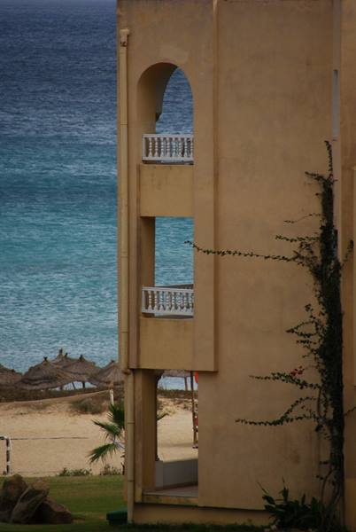 Ochre hotel facade with white balconies facing turquoise sea and thatched umbrellas