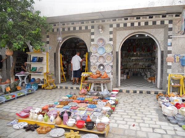Colorful pottery displayed before arched shops, a customer browsing plates and tagines