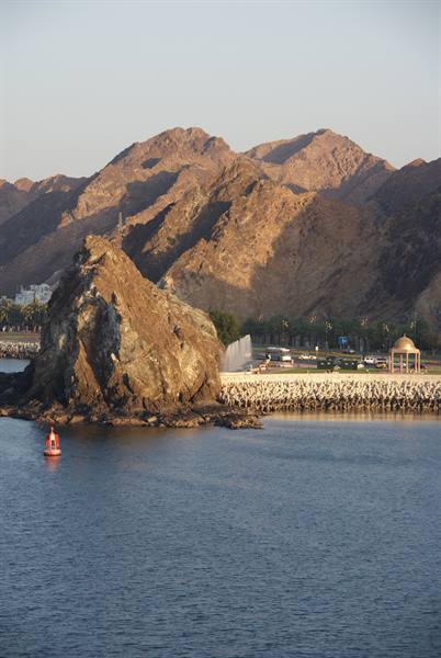 Rocky islet with red buoys, ochre mountains and corniche kiosk, Muscat