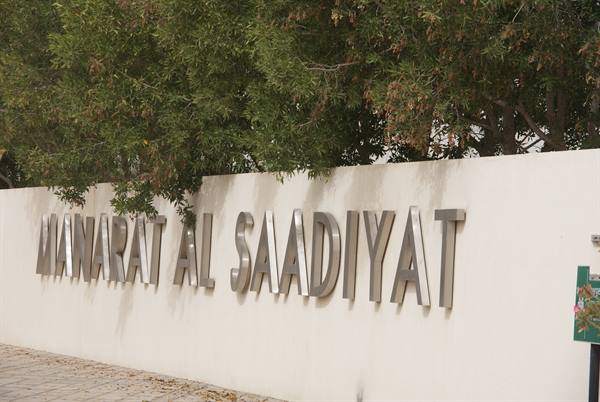 Metallic Manarat Al Saadiyat lettering on white wall, green tree, daylight