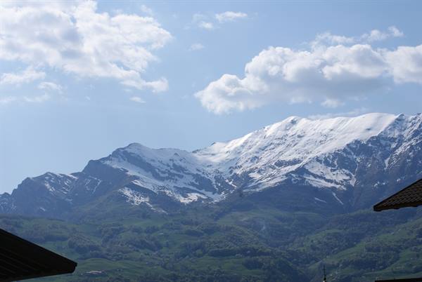 Snow-capped peaks above green hills, wooden rooftops in foreground under white clouds