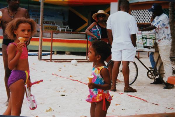 Two girls in swimsuits eat ice cream on sand, vendor with bicycle nearby