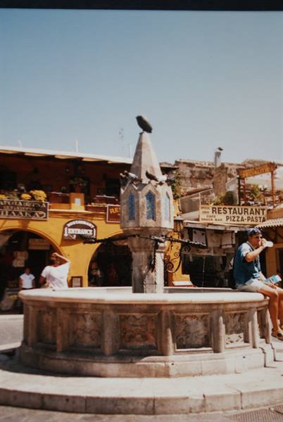 Medieval stone fountain with pigeons, tourists sitting beside restaurants