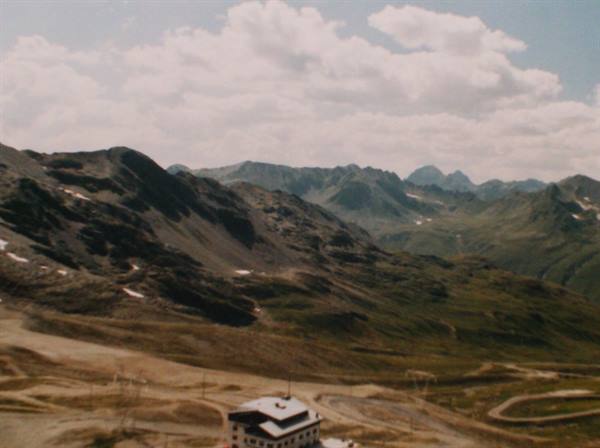 Stone mountain hut beneath rocky alpine peaks, white clouds and grassy slopes
