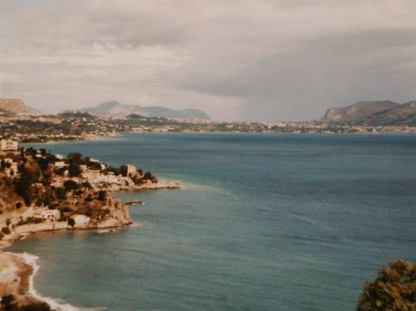 Turquoise bay with rocky shore, white houses and hills beneath grey clouds