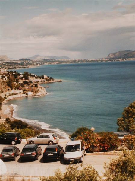 Cars parked above a sandy cove with turquoise sea and hills